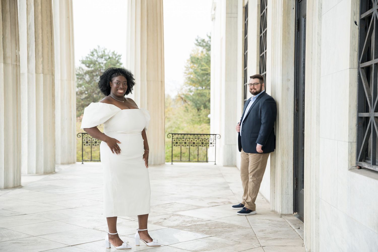 A couple stands apart on an elegant white columned porch during an outdoor photoshoot.