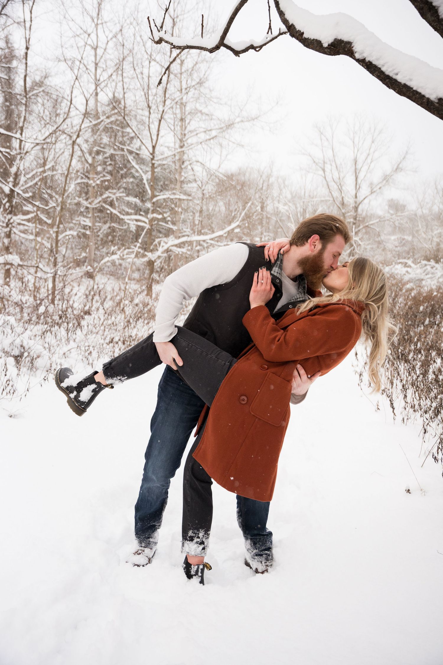 A couple shares a romantic winter embrace while standing in fresh snow surrounded by frost-covered trees.