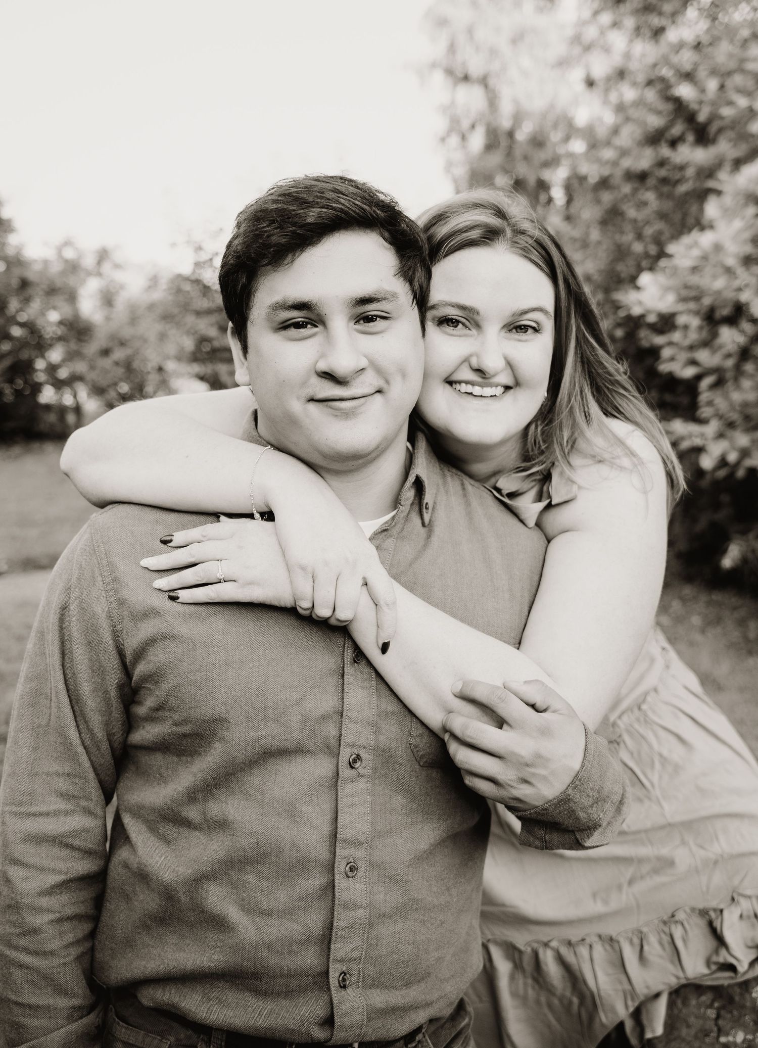 A romantic couple embracing outdoors in black and white, with the woman hugging from behind.