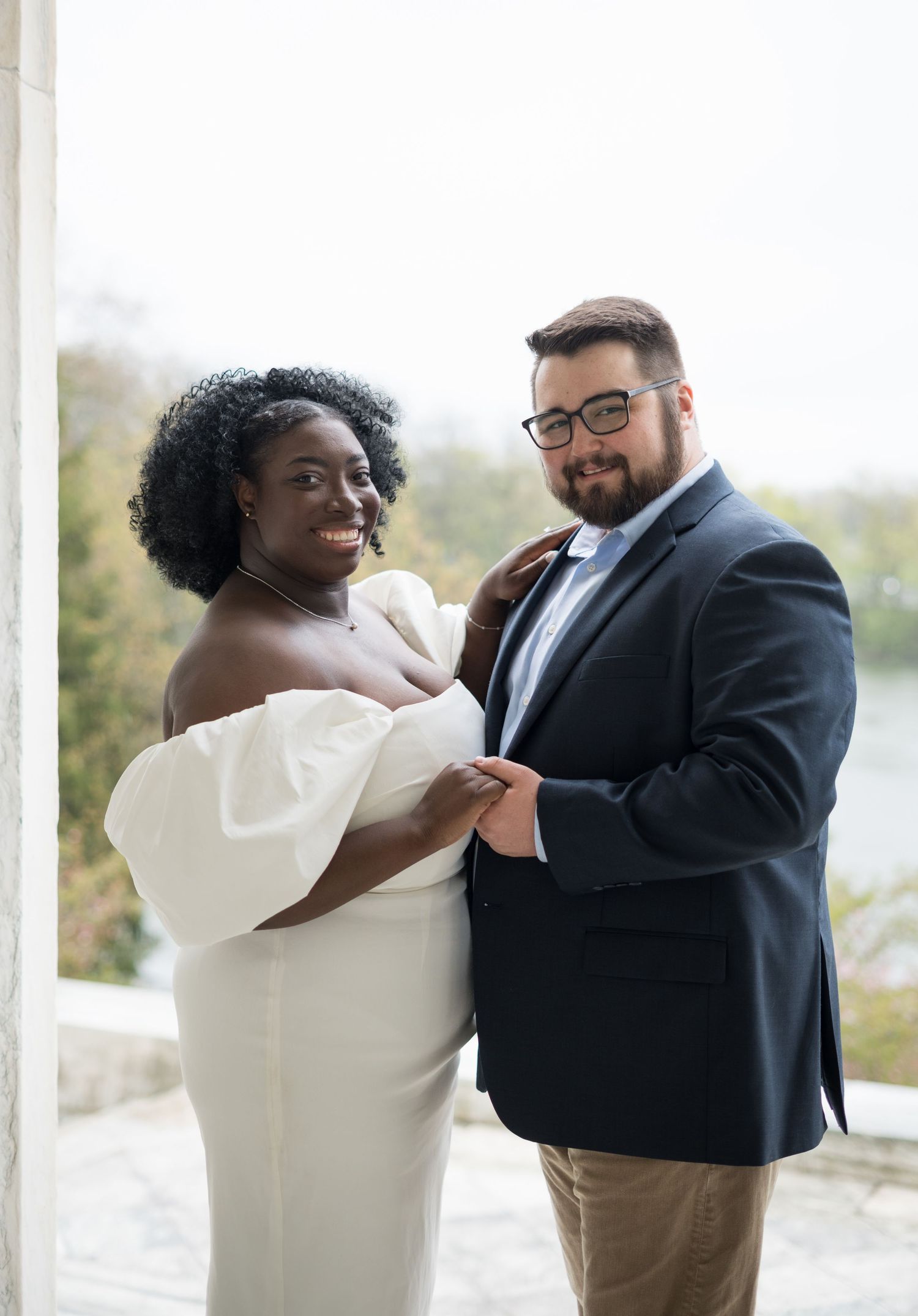 A couple in semi-formal attire stands together on a porch, with the person in white wearing an elegant off-shoulder dress.