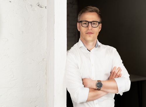 Professional portrait of person in white shirt and glasses leaning against white wall.