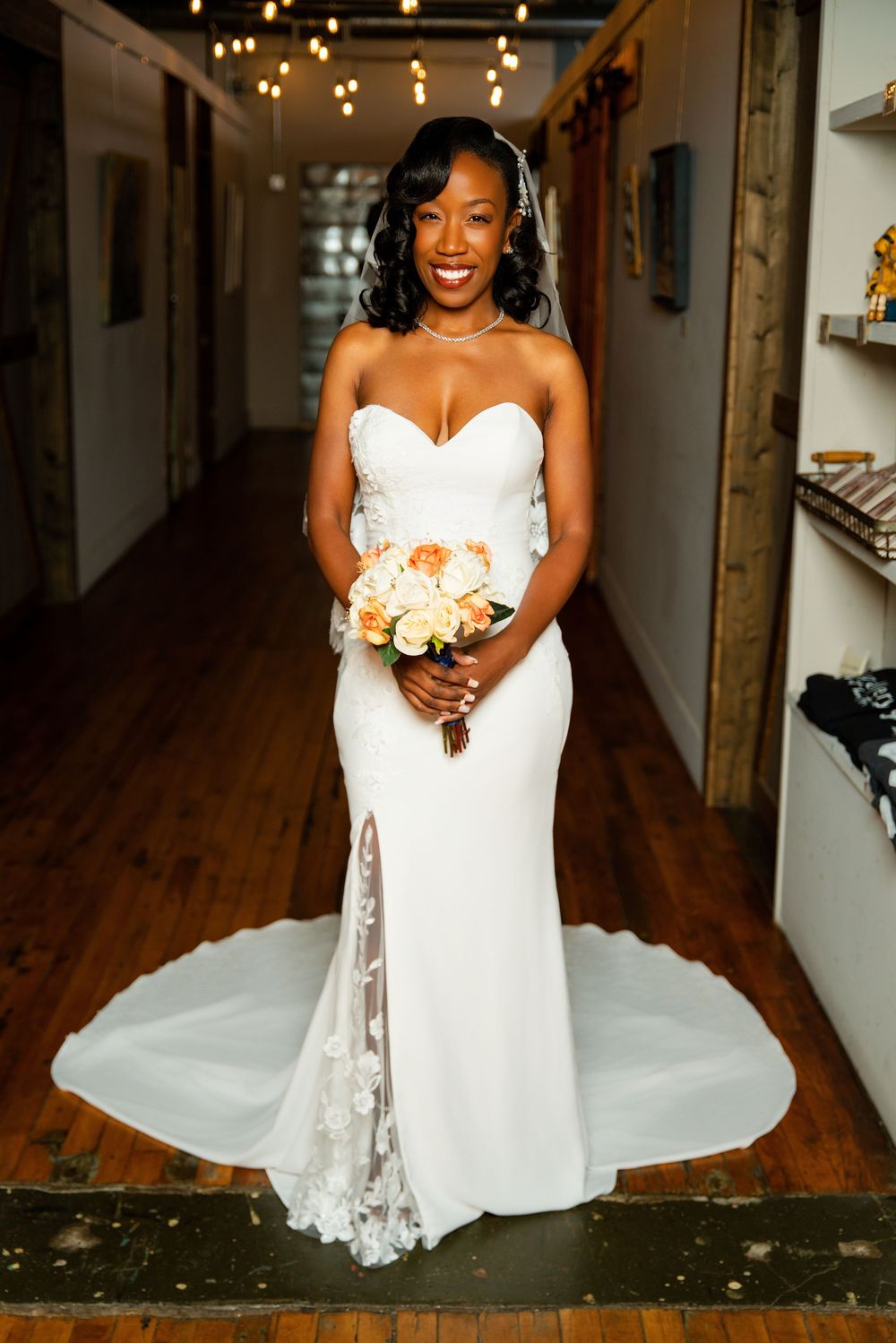 Beautiful bride in a strapless white lace wedding gown with sweetheart neckline holds peach and cream bouquet in rustic hallway.