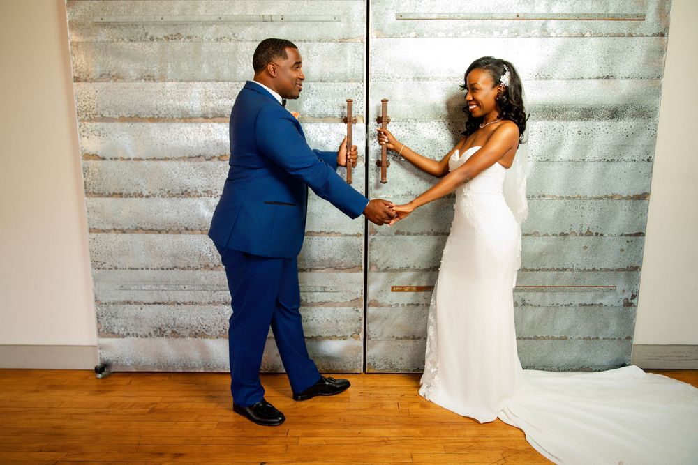 A couple in wedding attire shares a playful dance moment against a textured gray wall backdrop.