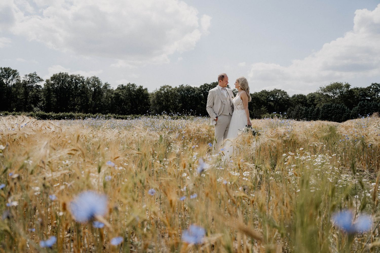 A couple in wedding attire stands together in a sunlit meadow filled with wildflowers and tall grass.