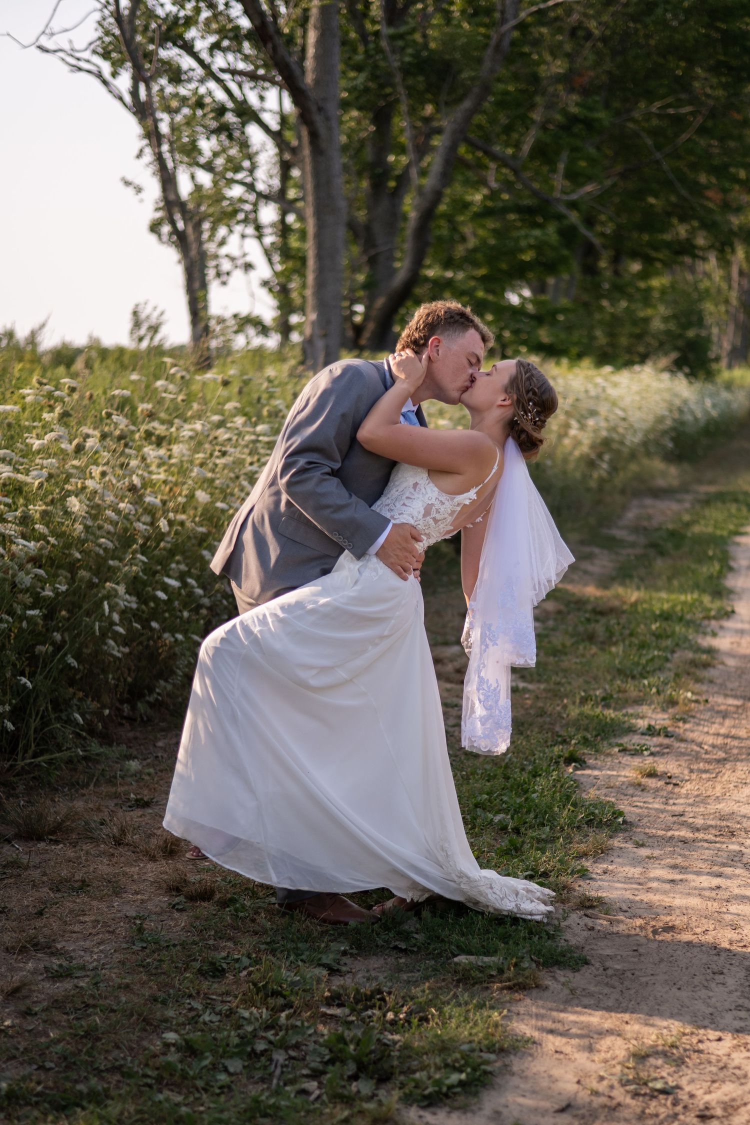 Elegant bride in lace gown poses in front of sunflower field at sunset.