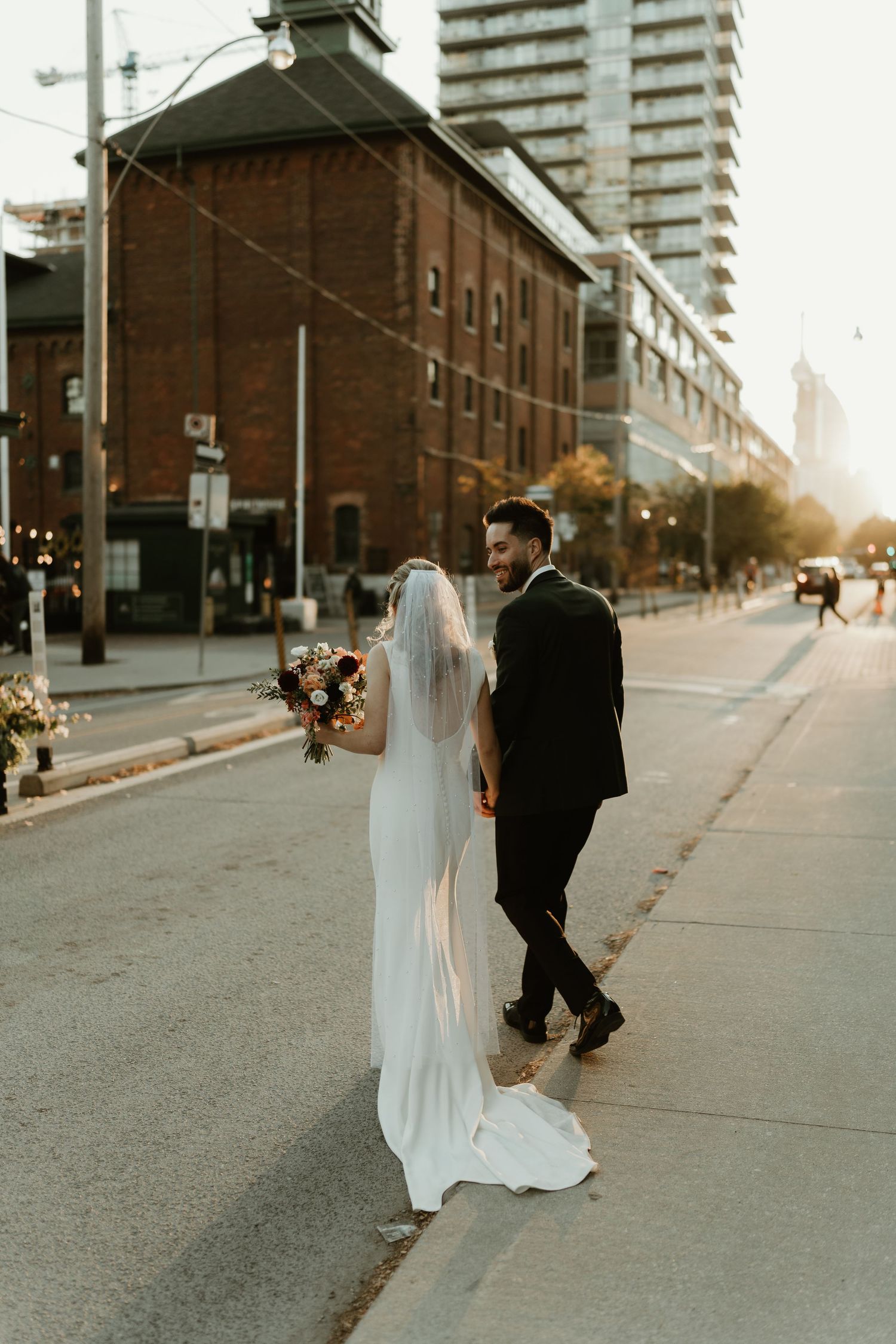 Bride and groom walking down a city street at sunset with urban architecture in background.