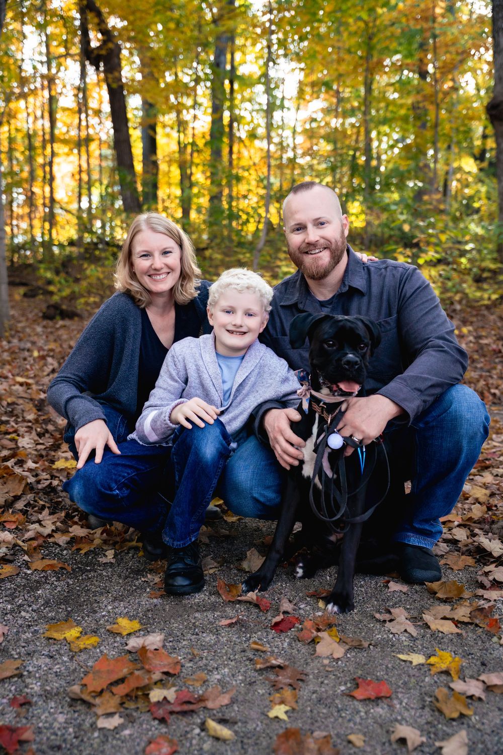 Family of two adults and a child pose with their black dog among colorful autumn leaves in a wooded park.