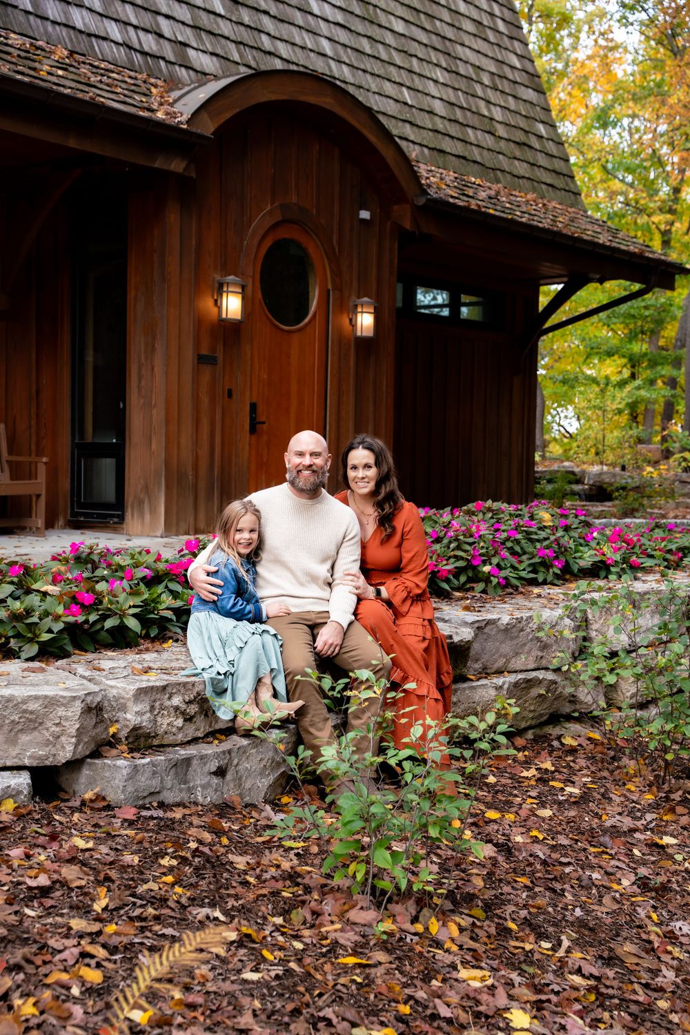 Family of three sitting on stone steps outside a rustic wood cabin surrounded by colorful fall foliage.