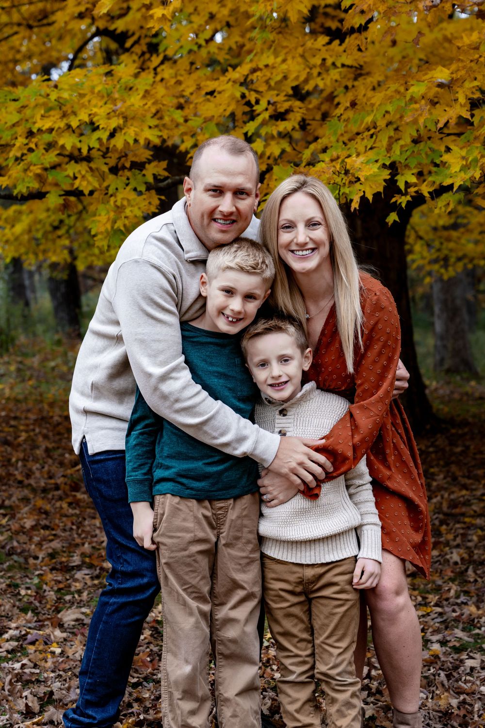 Family of four posing outdoors in autumn, parents hugging two young boys among golden fall foliage.