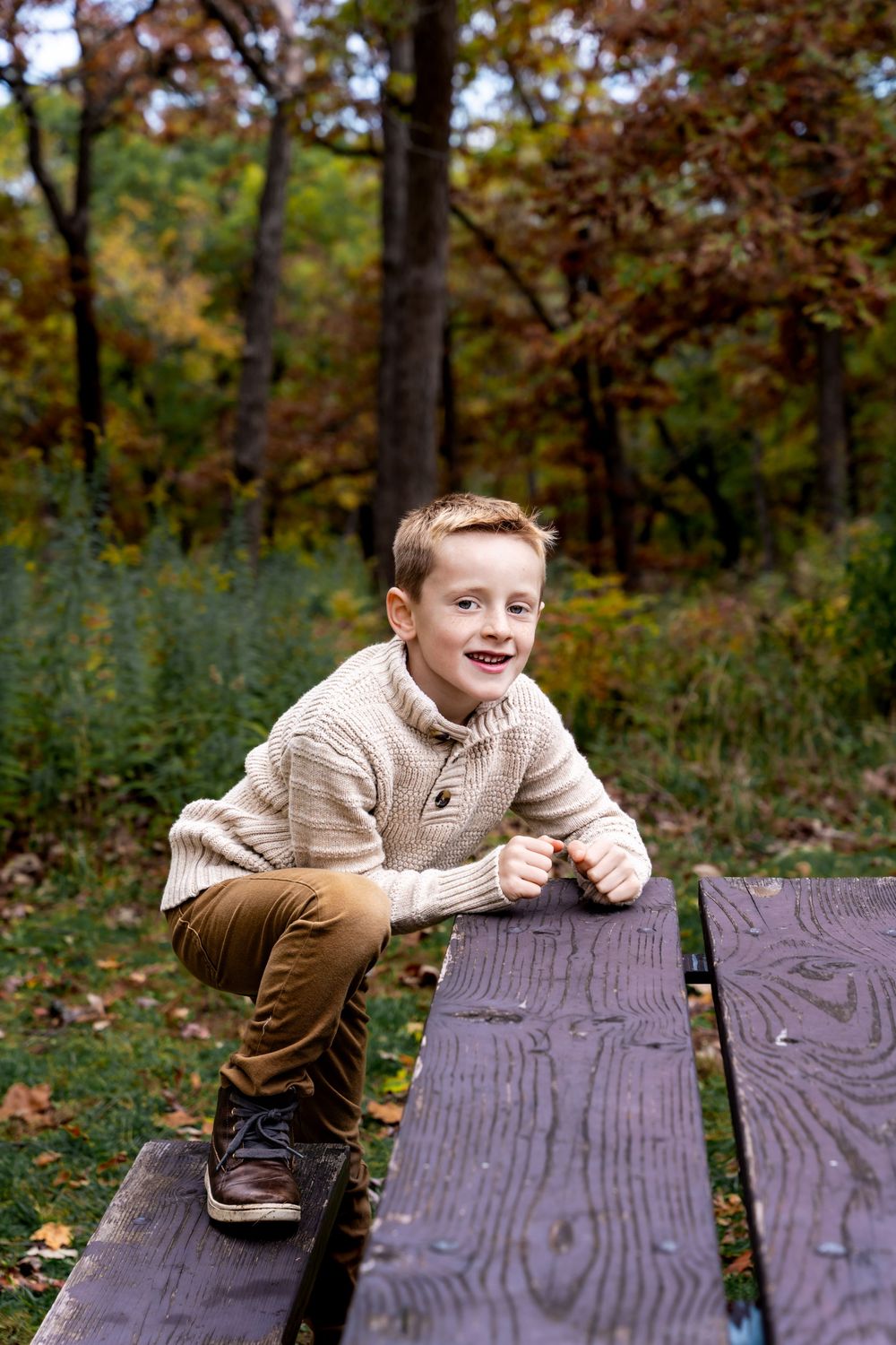 Smiling boy in beige sweater and brown pants crouching on a wooden picnic table in an autumn forest park.