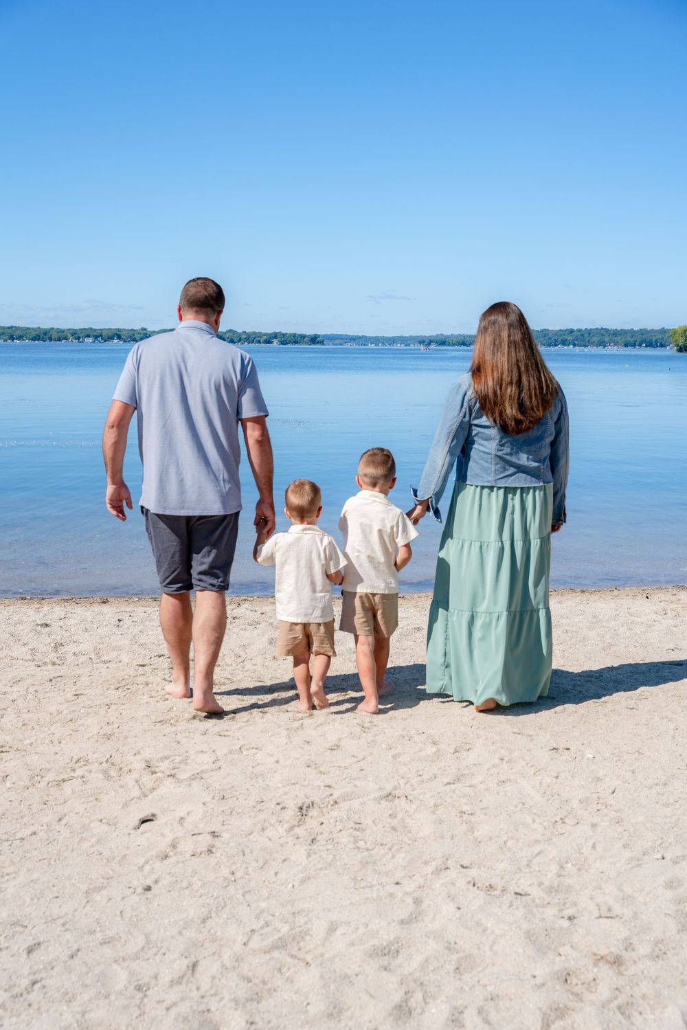 Family of four walking hand-in-hand on a sandy beach toward calm blue water on a sunny day.