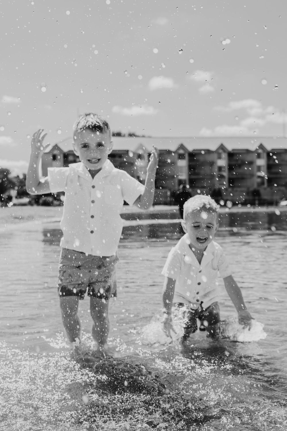Two young boys splashing and playing joyfully in shallow water near a beach resort in black and white photo.