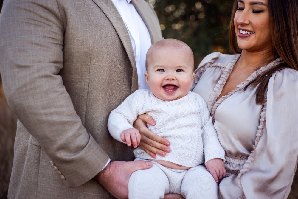 Laughing baby in white outfit held by parents outdoors.