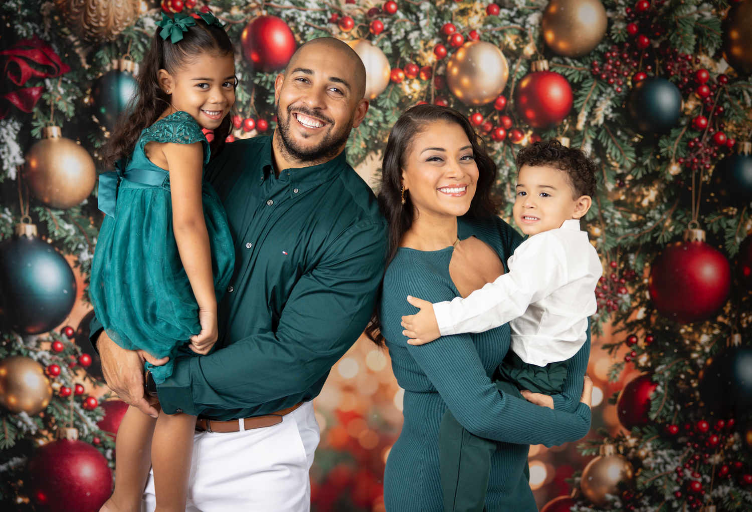 Family holiday portrait with coordinated outfits against Christmas ornament backdrop.