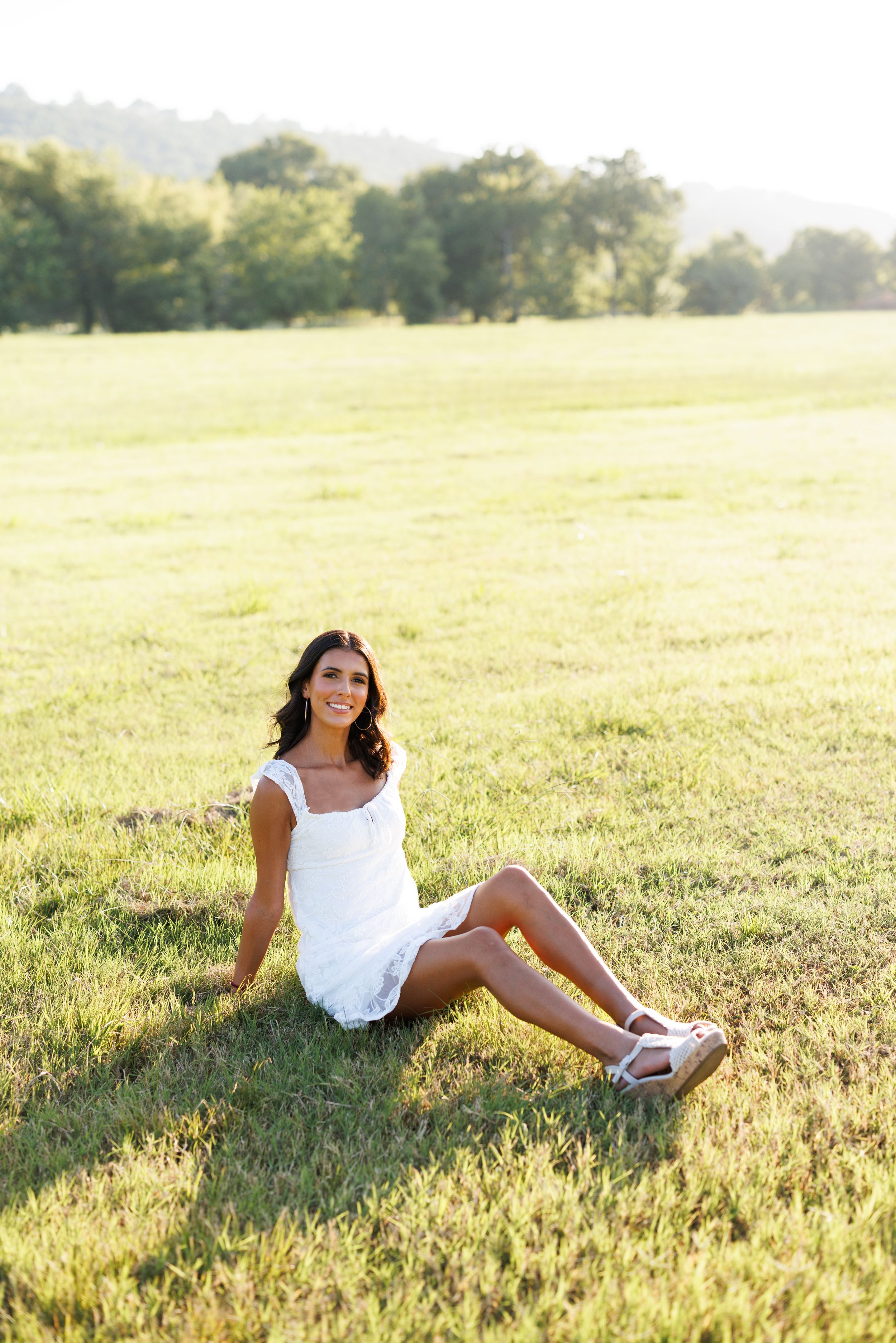 Summer portrait in white dress sitting on sunlit grassy field.