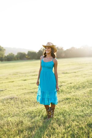 Woman in turquoise dress and cowboy boots standing in sunlit field.