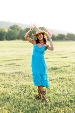 Playful pose in turquoise dress and cowboy hat with hands raised in sunny meadow.