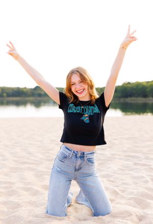 Person wearing black crop top and jeans poses joyfully on sandy beach at sunset.