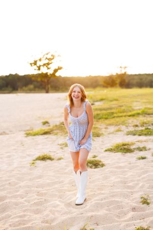 Person wearing blue top and white boots poses on sandy beach at sunset.