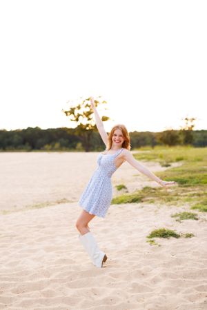 Person in blue floral dress leaps playfully on beach during sunset.