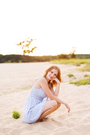 Person in blue floral dress sits gracefully on sandy beach at golden hour.
