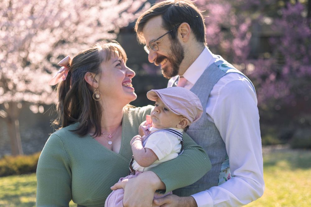 Happy couple smiling at their baby outdoors with pink cherry blossoms in the background on a sunny day.