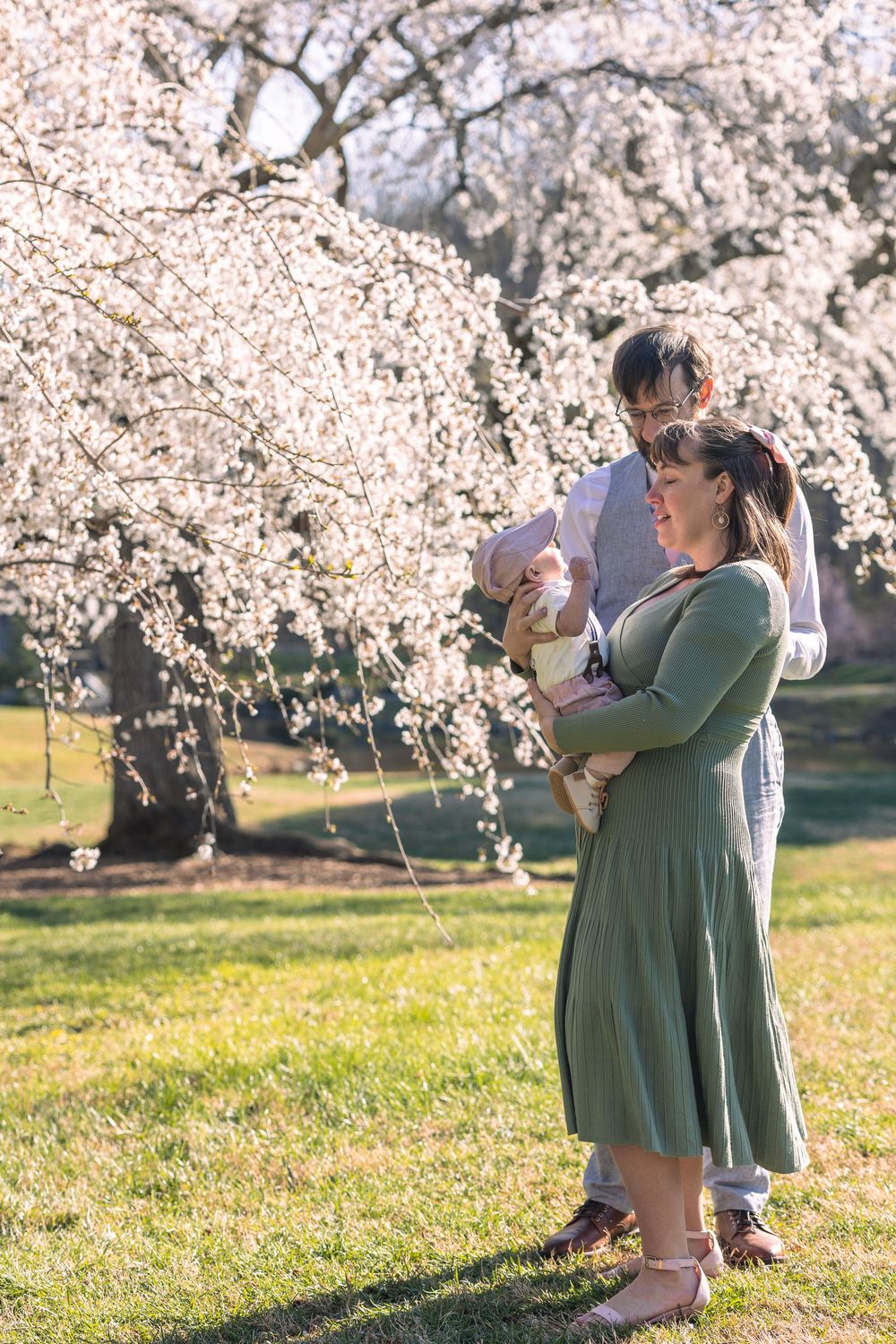 Couple holding newborn baby under blooming cherry blossom trees in spring park, woman in green dress.