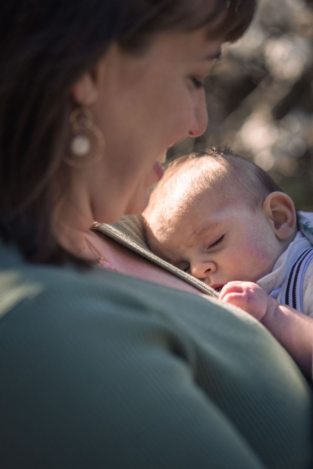 A mother tenderly gazes at her sleeping newborn baby cradled against her chest outdoors in soft sunlight.