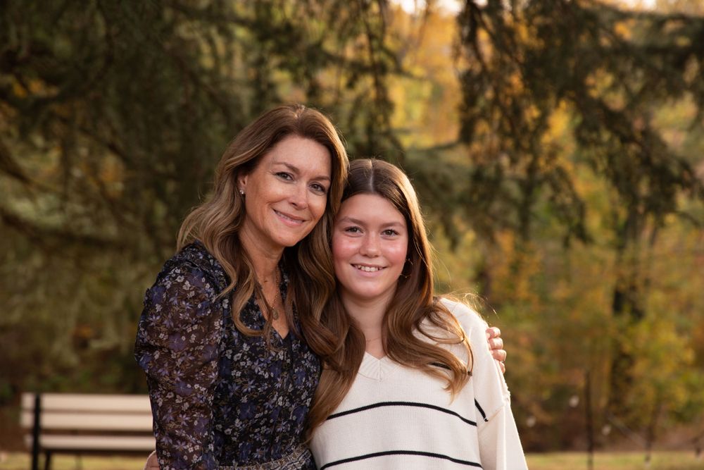 Two people share a warm embrace outdoors during autumn with trees in the background.