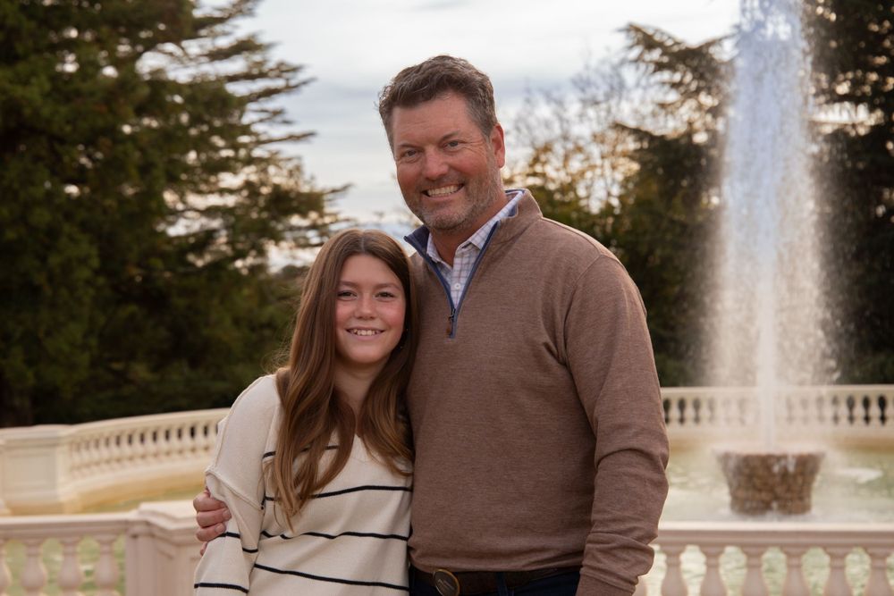 Two people smiling outdoors with a fountain and garden backdrop.