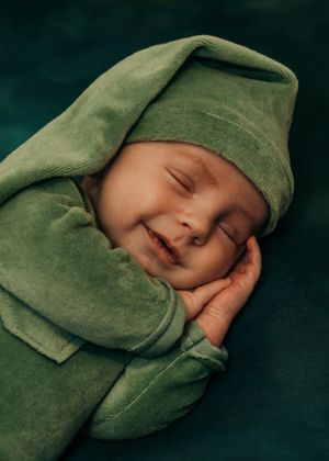 A baby wearing cozy green velvet clothing and matching hat sleeps peacefully on a dark background, during his newborn portrait session.