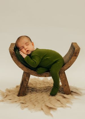 Professional portrait of sleeping newborn in green sleeper outfit rests peacefully on curved wooden prop with woven blanket underneath.