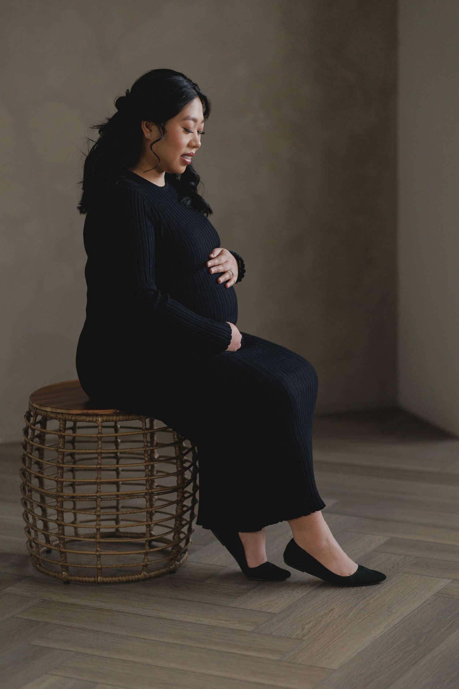 Side view of pregnant woman in black dress seated on woven stool against minimalist background.
