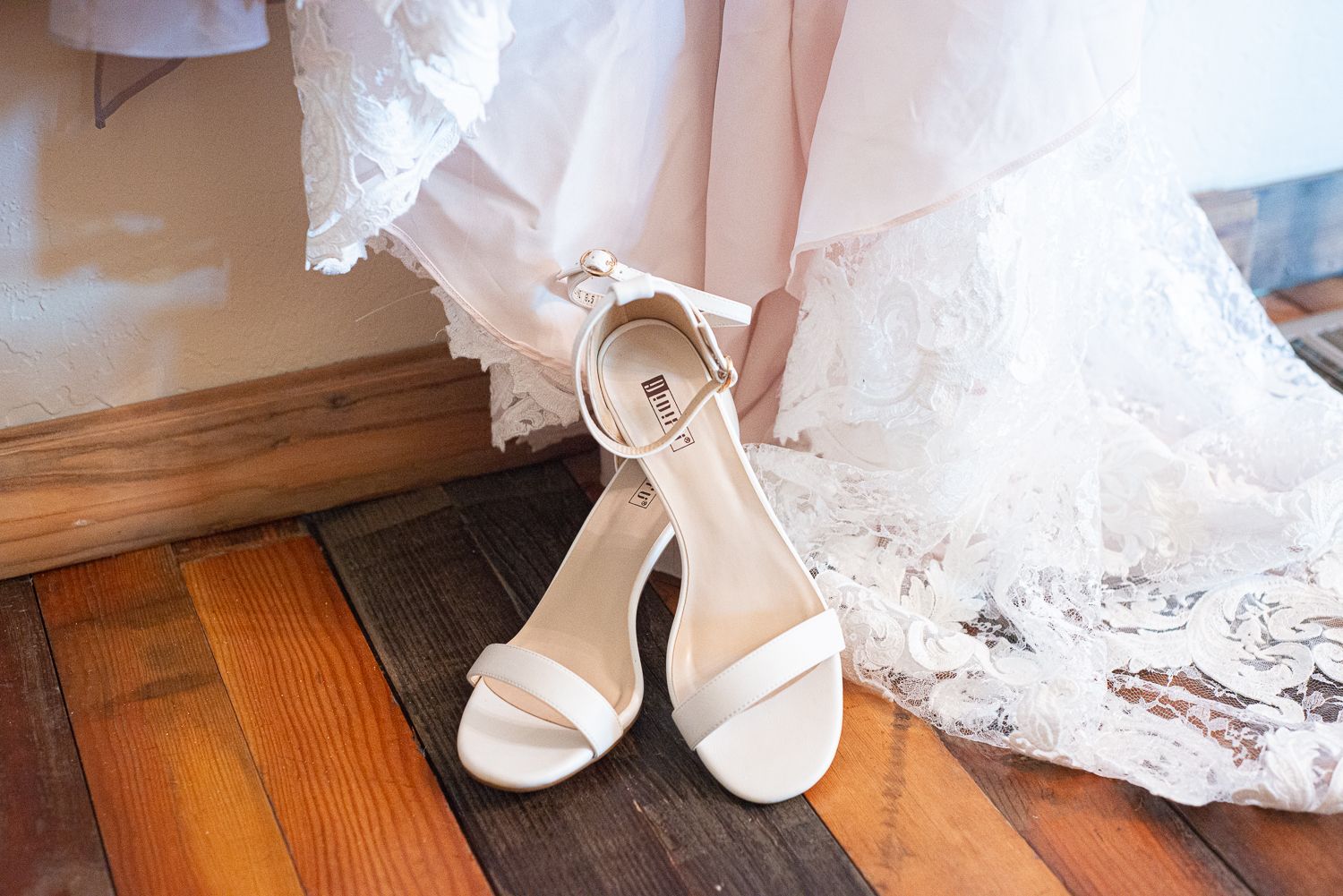 White bridal heels with ankle straps placed on a wooden floor next to flowing lace fabric.