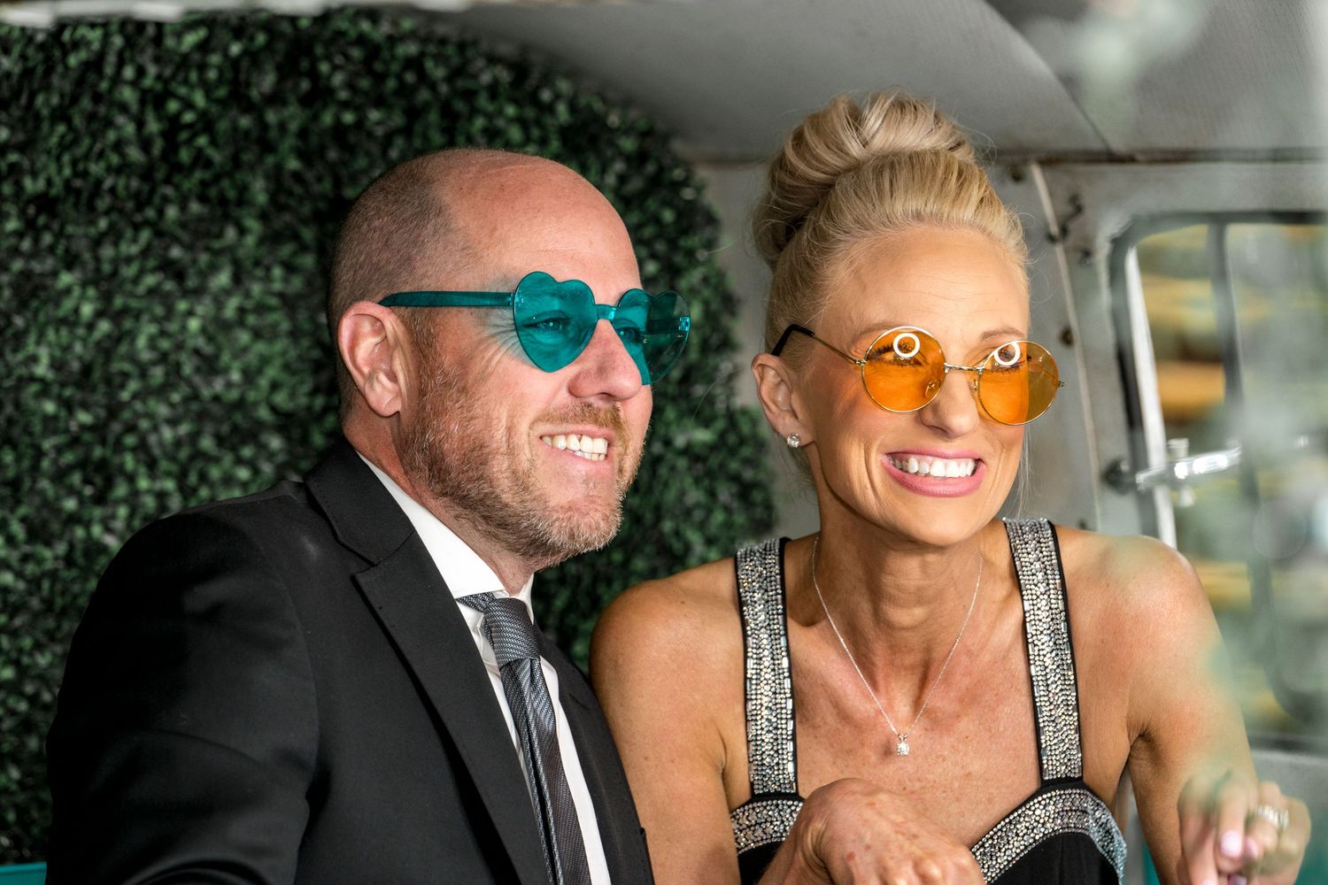 Couple wearing colorful sunglasses smiling in vehicle with greenery backdrop.