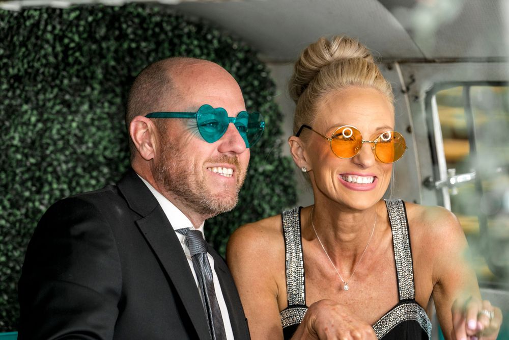 Couple wearing colorful sunglasses smiling in vehicle with greenery backdrop.