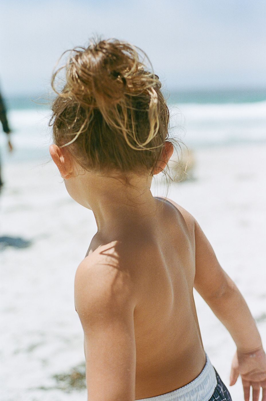 Back view of a child with messy blonde hair standing on a sunny beach.