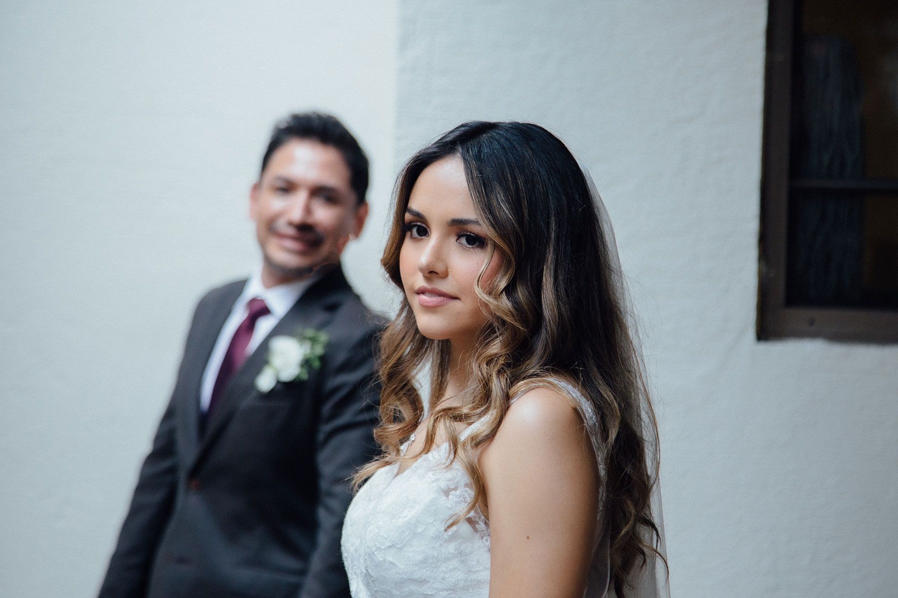 Bride in white lace dress glances back at groom in gray suit with boutonniere at outdoor wedding.