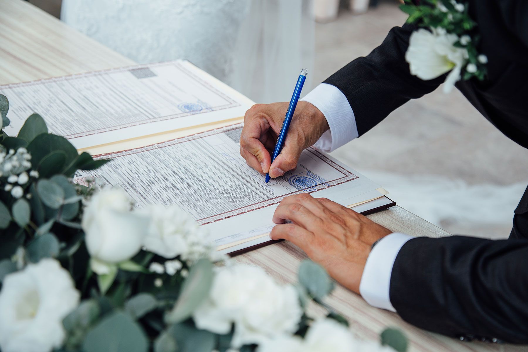 Groom signing wedding certificate with blue pen surrounded by white floral arrangements.