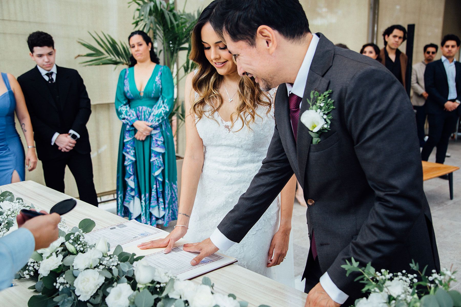 Bride and groom signing wedding register surrounded by white flowers and guests.