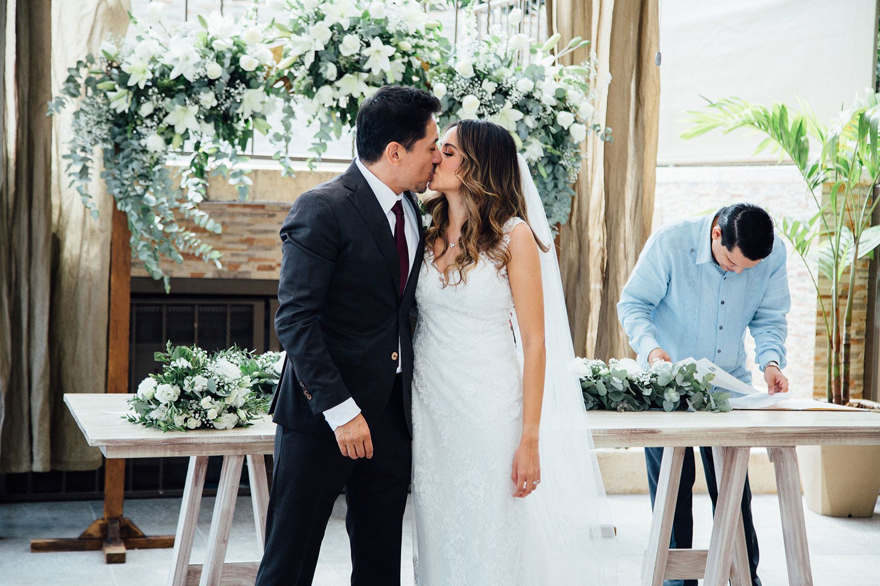 Newlyweds sharing first kiss at altar adorned with white floral arch decorations.