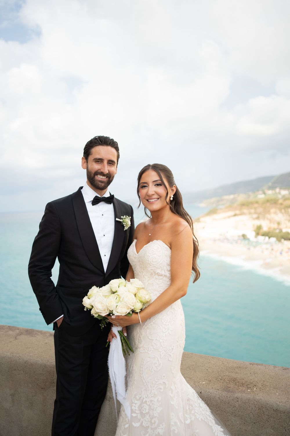 A wedding couple stands together against a beautiful coastal backdrop with white bouquet.