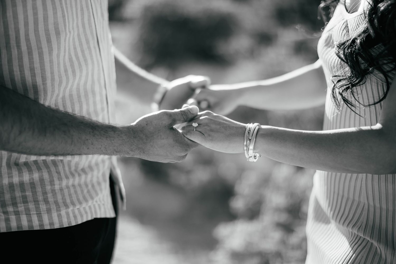 Close-up black and white photo showing hands with engagement rings against blurred background.