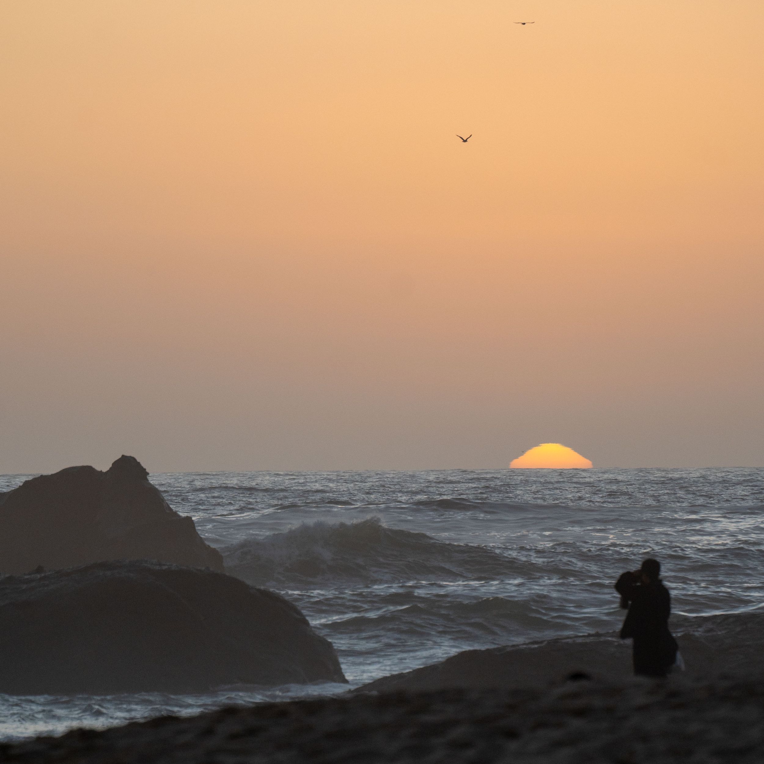 Silhouetted rocks against an orange sunset over the ocean.
