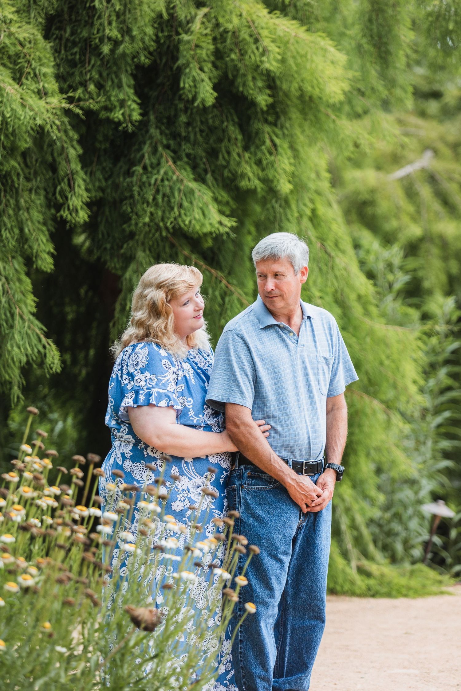 Couple shares tender moment on garden path surrounded by evergreen trees and flowers.