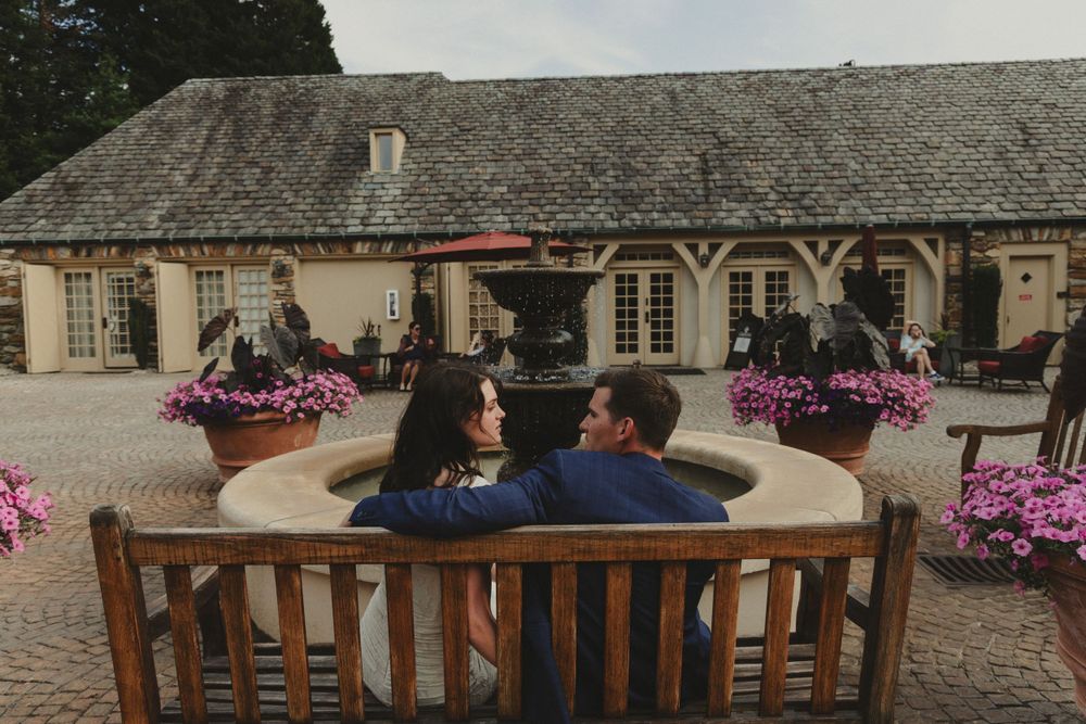 A couple relaxes on a wooden bench in front of a stone building with colorful flower displays.