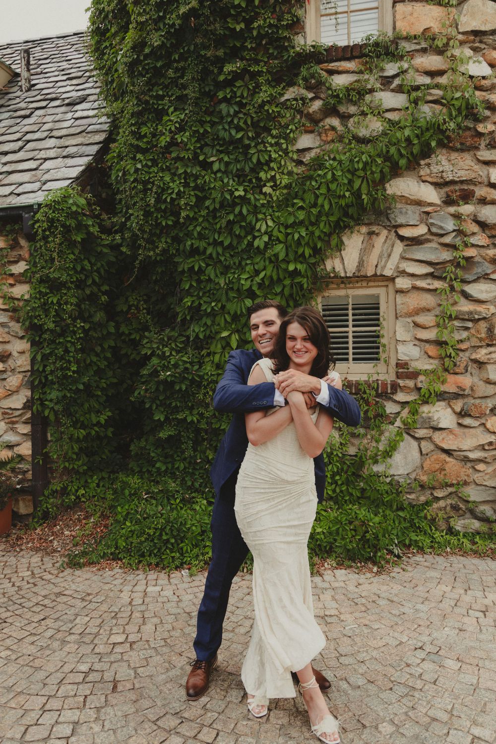 A couple embraces in front of a stone building covered in lush green ivy.