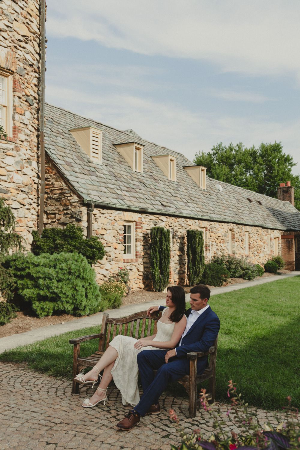 Couple relaxing on wooden bench against rustic stone building with ivy.