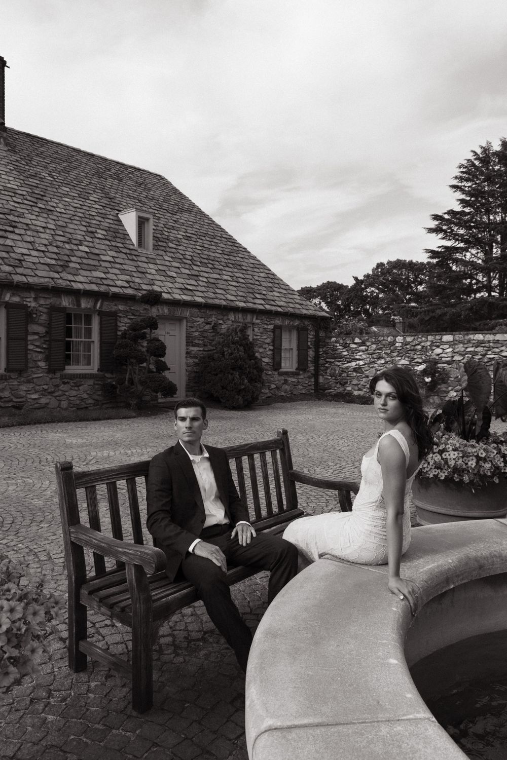 A black and white portrait of a couple in wedding attire seated by a curved stone wall near a cottage.