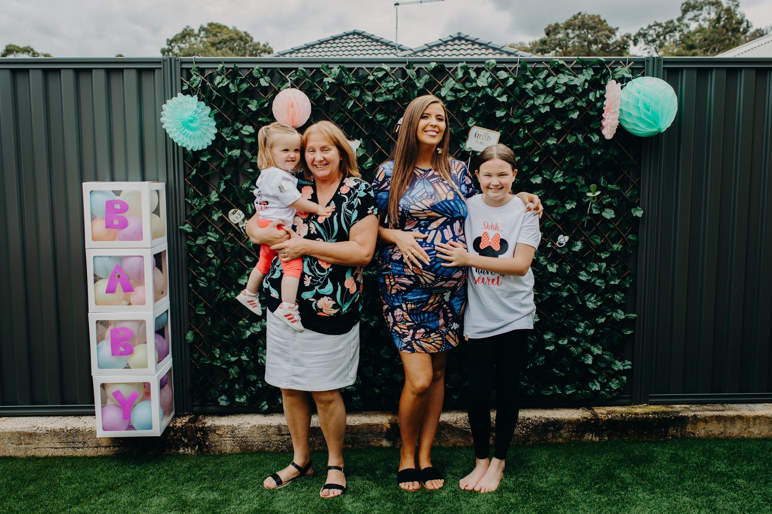 Party gathering with balloon decorations against green hedge wall backdrop.